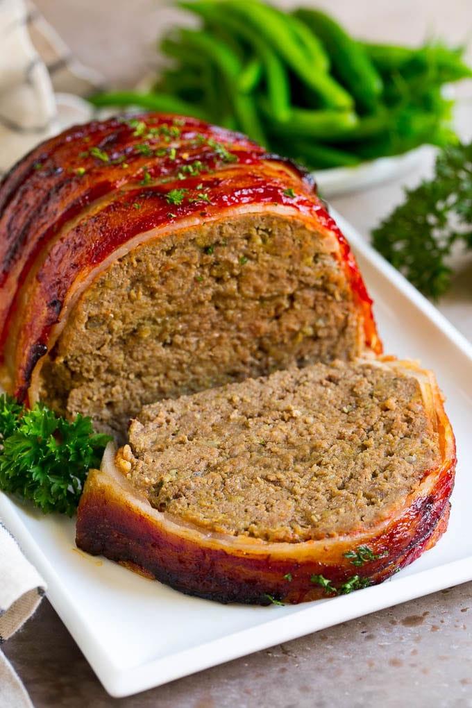 Hackbraten im Speckmantel Rezept | Rinderhackbraten #hackbraten #rindfleisch #rinderhack #speck #abendessen #abendessenimzoo Ein in Speck eingewickelter Hackbraten mit einer fehlenden Scheibe auf einer Servierplatte.