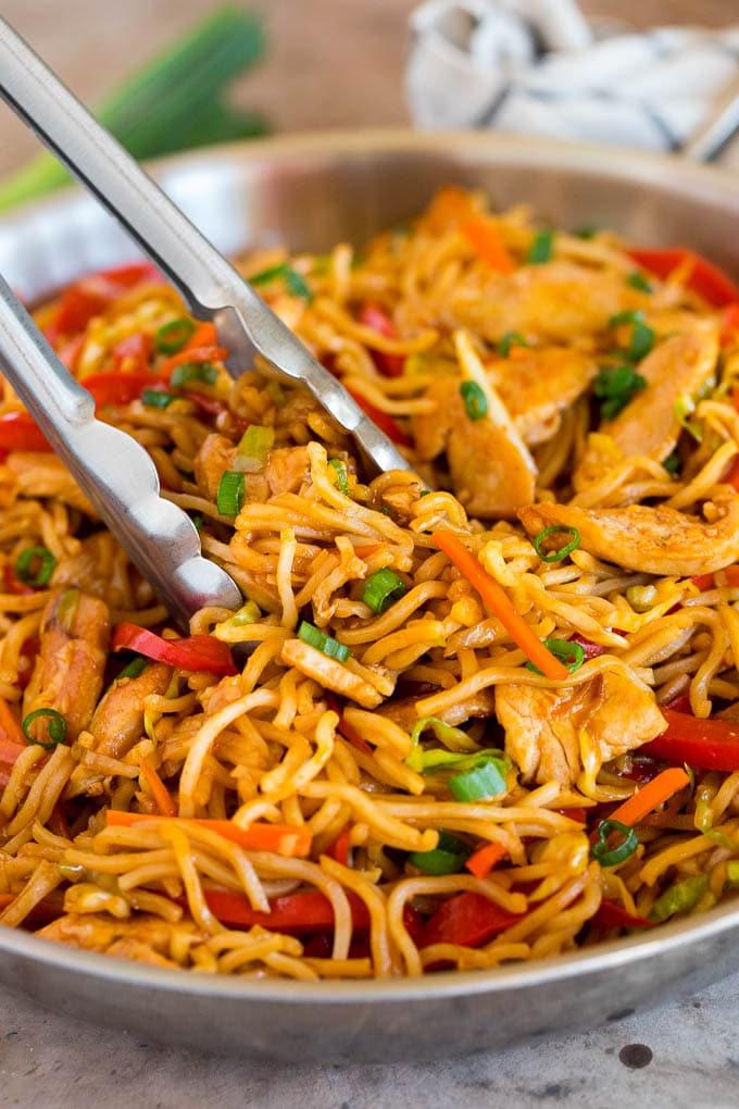 Diese Yakisoba Nudeln sind zarte, gebratene Nudeln, die in einer herzhaften Sauce mit Hähnchen und Gemüse zubereitet werden. Tongs serving up a portion of yakisoba noodles with vegetables and chicken.