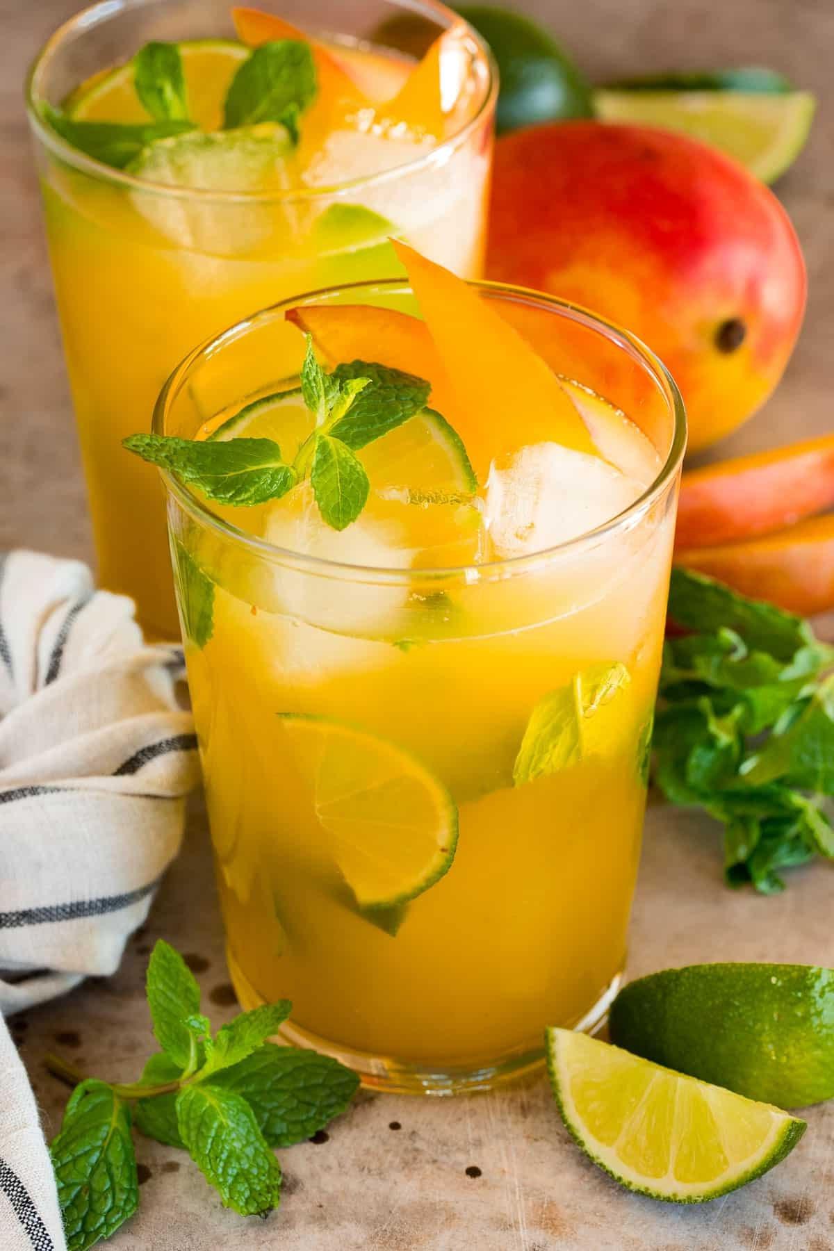 Mango Mojito 2 Gläser mit Mango Mojito, garniert mit frischer Minze, Limettenscheiben und saftigen Mangoscheiben. Einladendes, kühles Sommergetränk für heiße Tage.