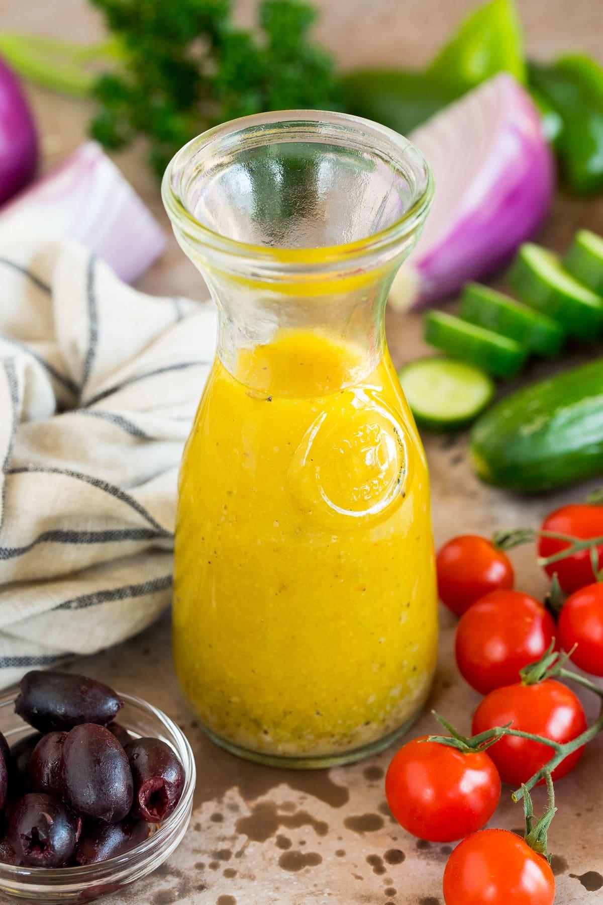 Griechisches Salatdressing 2 Eine Flasche griechisches Salatdressing, umgeben von frischem Gemüse wie Gurken, Tomaten und Zitronen.