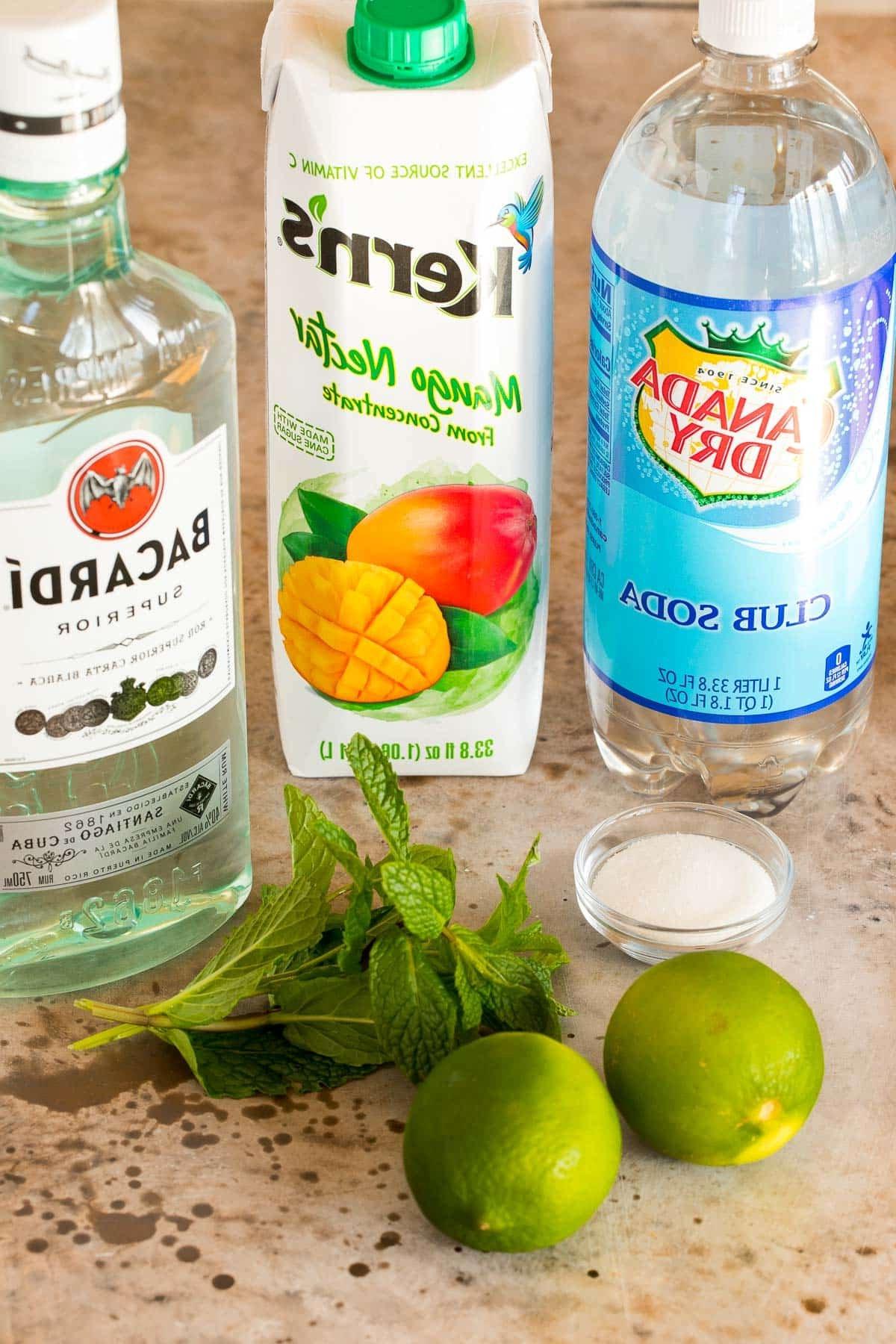 Mango Mojito 3 Flaschen mit weißem Rum, Mangonektar und Sodawasser, dazu frische Limetten, Zucker und Minze auf einem rustikalen Holztisch. Alle Zutaten bereit zum Mixen des tropischen Mojitos.