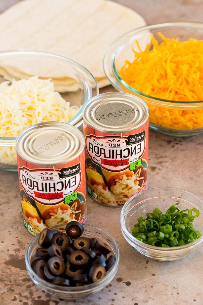 Dosen mit Enchilada-Soße und Schalen mit geriebenem Käse, grünen Zwiebeln und Oliven – alle Zutaten vorbereitet.