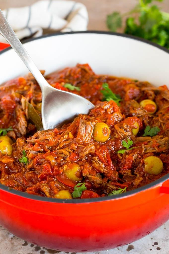 Dieses Ropa Vieja Rezept ist ein traditionelles Gericht aus Rinderbraten, das mit Tomaten und Gewürzen zart geschmort, dann zerzupft und mit Oliven serviert wird. Ein Servierlöffel in einem Topf Ropa Vieja mit Koriander garniert.