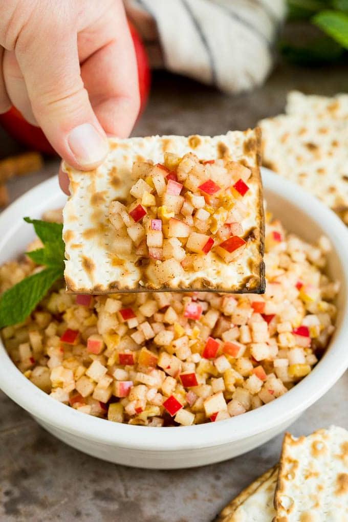 Dieses Charoset Rezept ist eine Mischung aus Äpfeln, Walnüssen und Honig, die zusammen eine süße Paste bilden, die ein wesentlicher Bestandteil jeder Passah-Mahlzeit ist. Eine Hand hält ein Stück Matze mit einer Portion Charoset darauf.