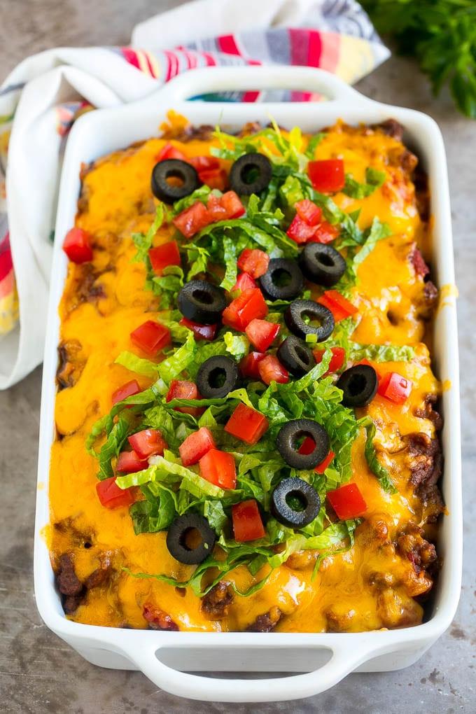 Ein Taco Auflauf in einer Auflaufform, bedeckt mit geschmolzenem Käse, frischem Salat, Tomatenwürfeln und Oliven.