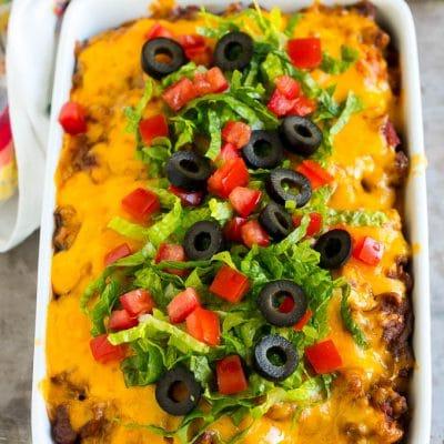 Ein Taco Auflauf in einer Auflaufform, bedeckt mit geschmolzenem Käse, frischem Salat, Tomatenwürfeln und Oliven.