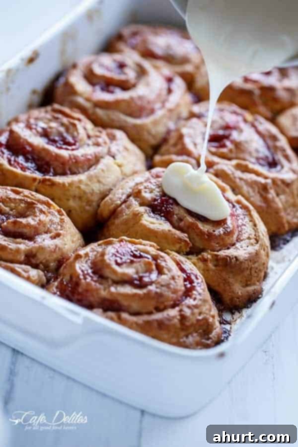 Creamy white frosting being poured over warm No Yeast Strawberries And Cream Cinnamon Rolls