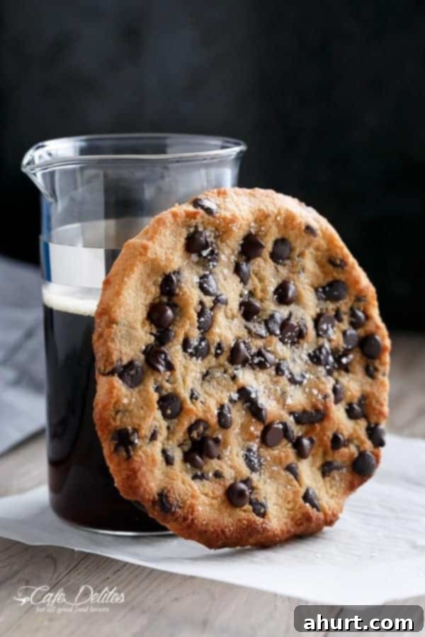A freshly baked single serve jumbo low carb chocolate chip cookie on a white plate with a cup of black coffee, highlighting its golden edges and melted chocolate chips.