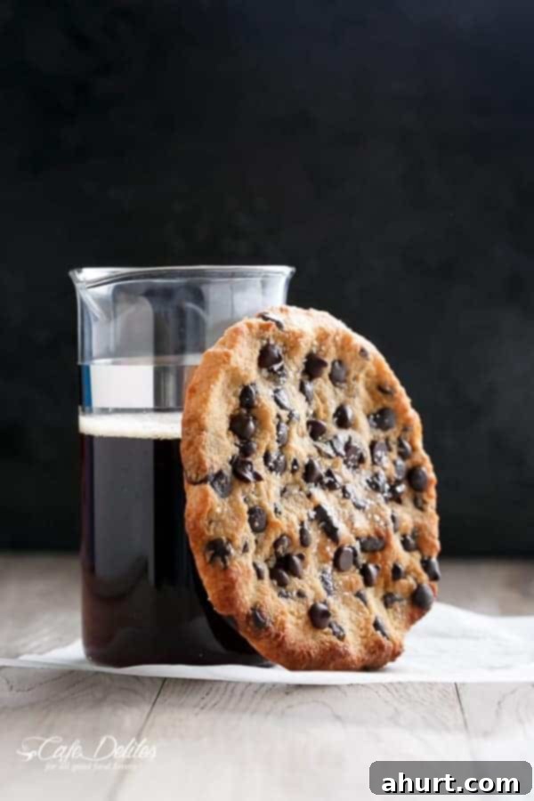 A single serve jumbo low carb chocolate chip cookie resting next to a refreshing cup of black coffee, suggesting a perfect pairing for a quiet treat moment.