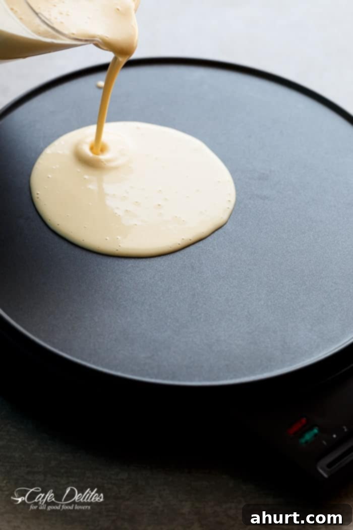 Stream of eggnog crêpe batter being poured onto a hot crêpe griddle, with a utensil spreading it evenly