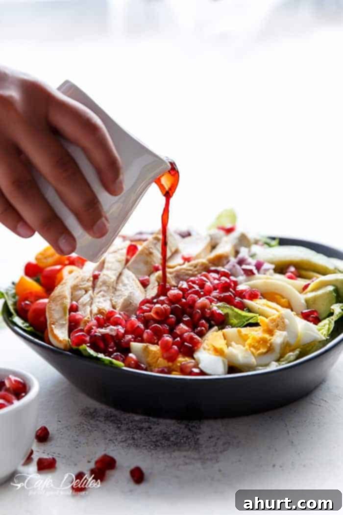 Jeweled Cobb Salad 6 Red wine vinaigrette being drizzled from a jug over a vibrant Pomegranate Chicken Bacon Avocado Salad, making the ingredients glisten.