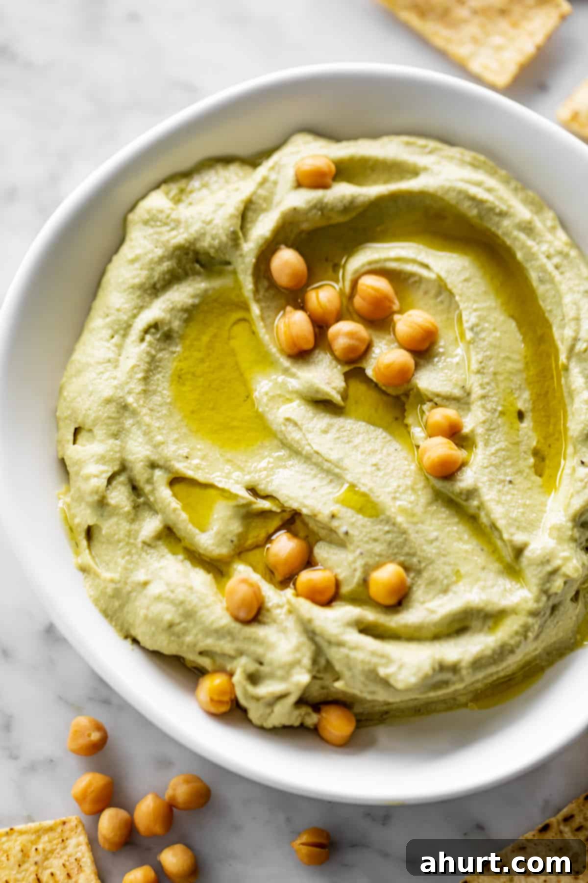 Creamy Avocado Hummus 2 Avocado Hummus served in a white bowl, drizzled with olive oil and topped with whole chickpeas and paprika. Corn chips are scattered on the table.