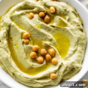 Creamy Avocado Hummus 11 Avocado Hummus served in a white bowl, drizzled with olive oil and topped with whole chickpeas and paprika. Corn chips are scattered on the table.