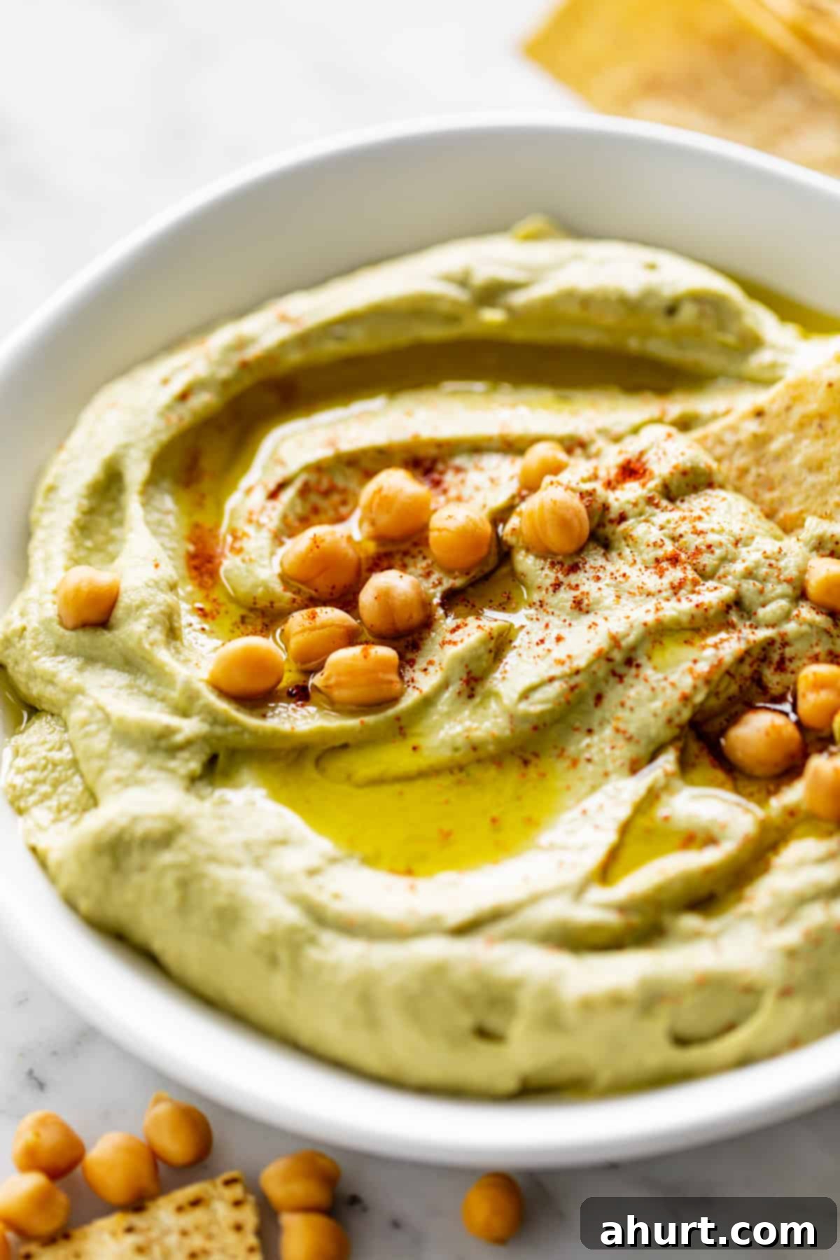 Creamy Avocado Hummus 10 Avocado Hummus served in a white bowl, drizzled with olive oil and topped with whole chickpeas and paprika. Corn chips are scattered on the table.