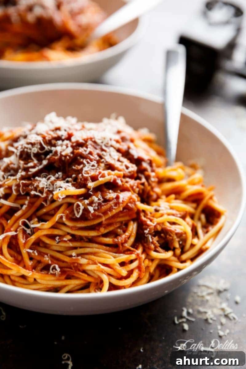 Fall-Apart Tender Slow Cooker Beef Ragu 2 A serving of Slow Cooker Beef Ragu in a white bowl, garnished with fresh basil leaves, with a spoon ready to serve. The sauce is rich and thick, showcasing tender shredded beef.