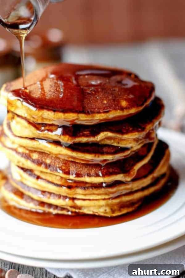 Tall stack of golden brown Caramel Chip Pumpkin Pancakes drizzled with maple syrup and caramel sauce, garnished with a few loose caramel chips on top, set on a rustic plate.