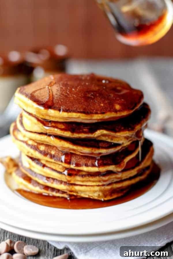 Side view of a stack of fluffy Caramel Chip Pumpkin Pancakes with melted caramel chips visible, drenched in maple syrup, served on a white plate with a fork.