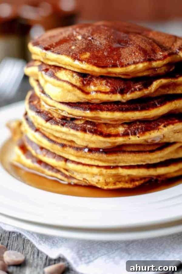 Side view of a serving of three Caramel Chip Pumpkin Pancakes on a white plate, generously topped with maple syrup and a dollop of whipped cream, ready to be enjoyed.