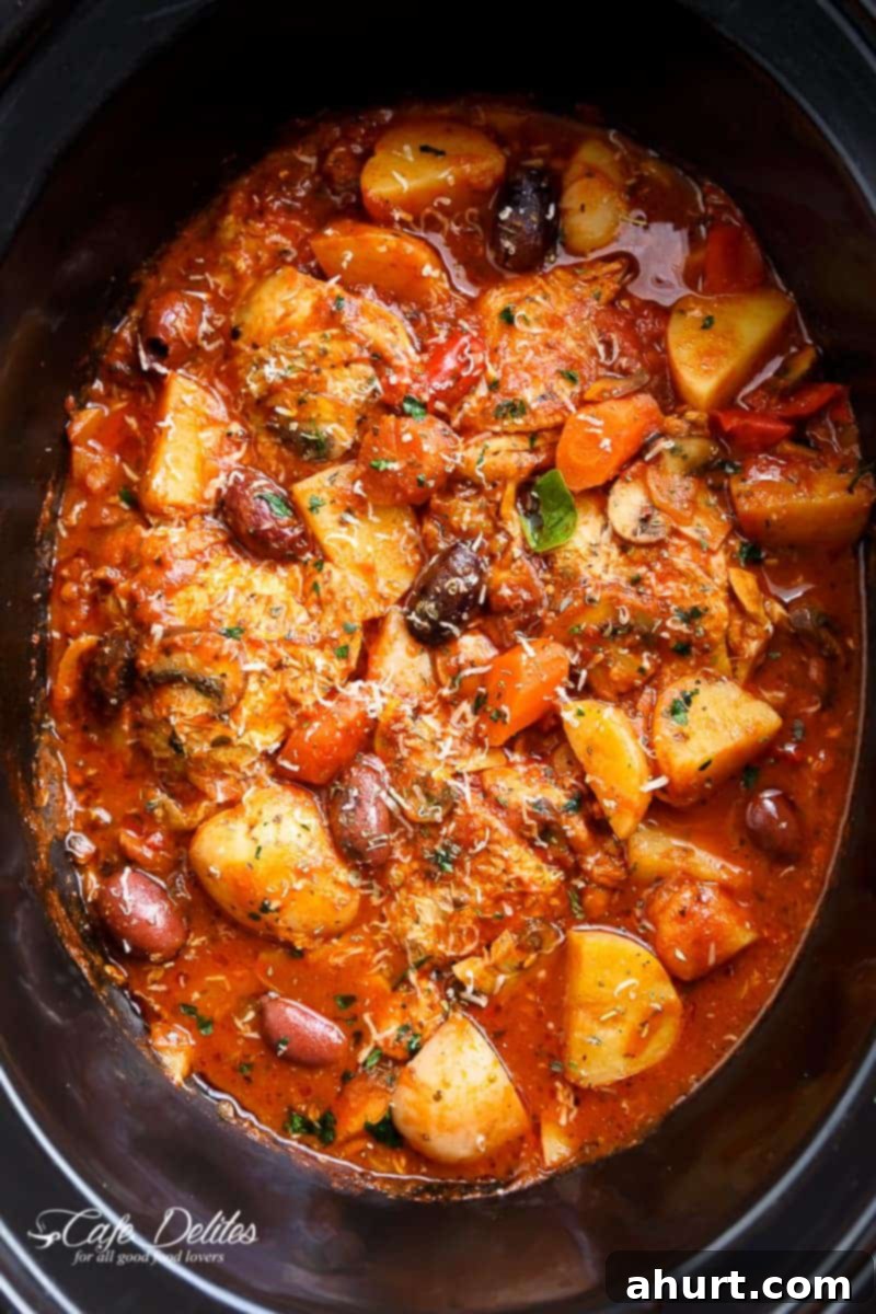 Set It and Forget It Chicken Cacciatore 3 Top view image of A rich, red stew full of chicken, roughly chopped vegetables being stirred by a white spatula in a slow cooker.