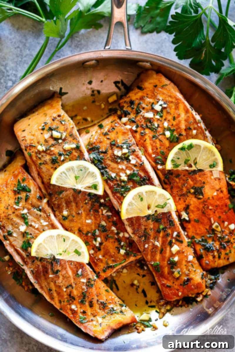 Four pieces of salmon in a pan with cooked herbs, garlic, melting butter and lemon slices on top.