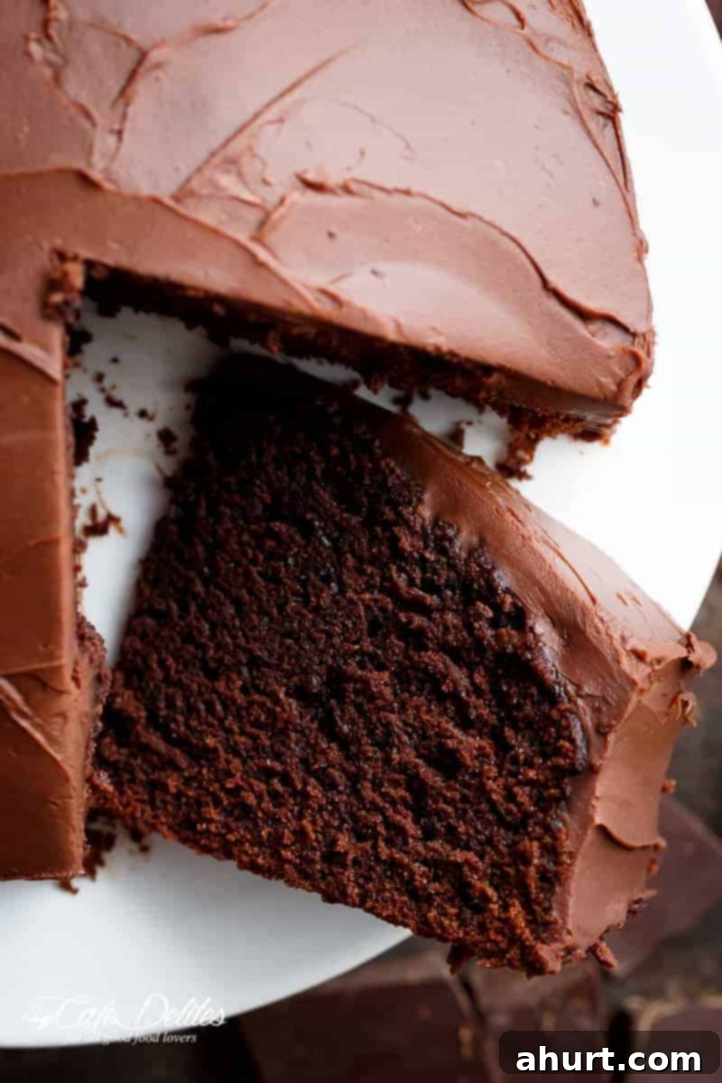 Decadently Fudgy Chocolate Cake 6 A rich fudgy chocolate cake, frosted in a decadent chocolate frosting, is shown sitting on a white plate. The first slice of cake has been cut lifted up from the rest of the cake. The chocolate frosting is smooth and shiny, while the cake is dense and moist looking. A pieces of chopped, chocolate cake is laying white plate to display the moist fluffy inside.