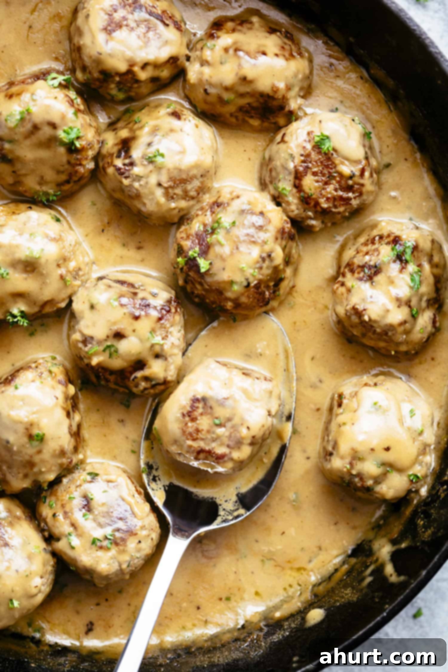 A top-down view of a cast iron skillet brimming with beautifully browned Swedish meatballs, generously coated in a rich, creamy, beige gravy, speckled with fresh herbs for garnish. The skillet rests on a dark, textured surface, emphasizing the warmth and rustic charm of the dish.