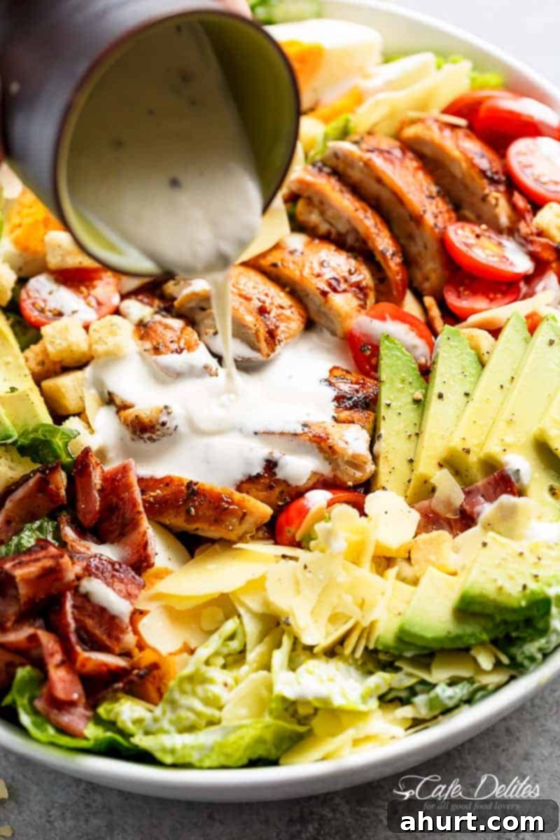 A close-up shot of creamy Caesar dressing being drizzled generously over a fresh Grilled Chicken Cobb Caesar Salad.