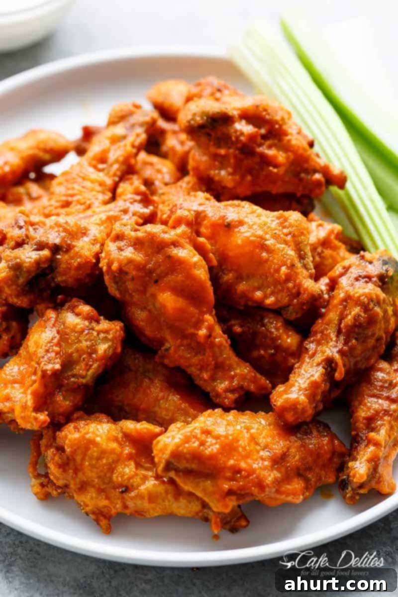 Golden Crisp Baked Buffalo Wings 2 The image showcases a delicious pile of chicken wings coated in a vibrant orange buffalo sauce. The wings are piled high on a white plate, their glistening skin reflecting light and hinting at their crispy texture. Accompanying the wings are several sticks of fresh celery, providing a cool and crunchy contrast to the spicy wings. The background is intentionally blurred, further emphasizing the chicken wings as the star of the show.