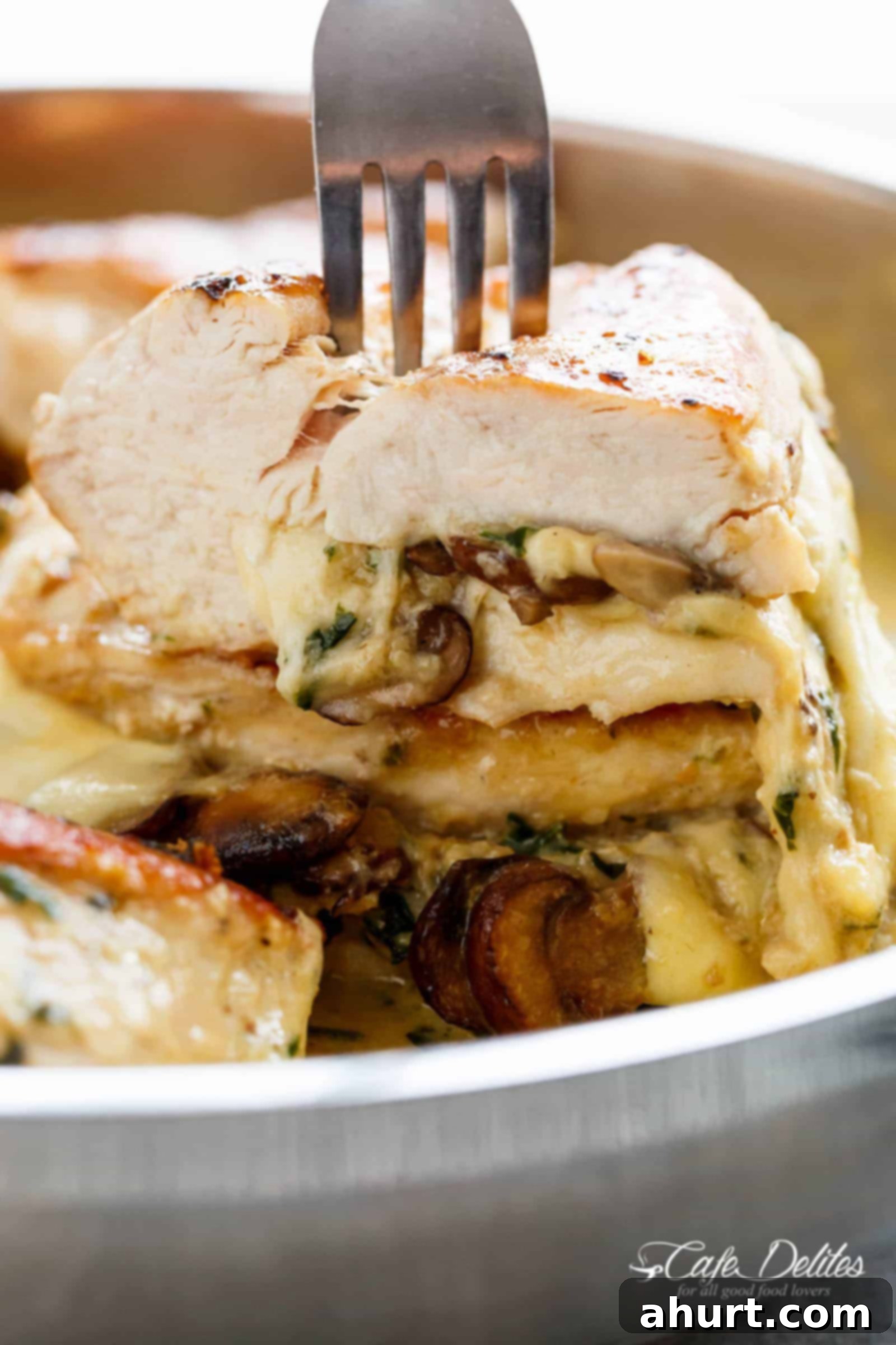 A fork pierces through cheesy mushroom stuffed chicken cut open! The piece is stacked on another stuffed chicken breast WITH the optional cream sauce in a silver frying pan.