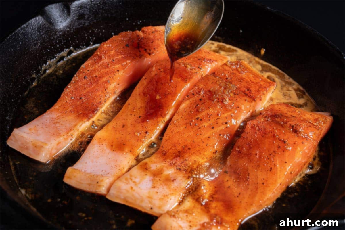 Glazed Honey Garlic Salmon 6 An image of salmon being cooked in a skillet with sauce. A spoonful of soy sauce is being added to the fillets as they cook.