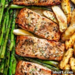 Decadent Garlic Butter Salmon 11 The image shows four salmon fillets on a baking sheet, along with asparagus spears and potato wedges. The salmon is covered in a brown glaze and garnished with fresh herbs and lemon slices. The bright green baked asparagus is soft and the potatoes are slightly browned after baked