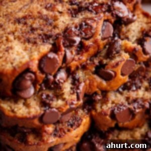 The Ultimate Chocolate Chip Banana Bread 11 freshly baked banana bread has a golden brown crust and the inside is a pale yellow color. The bread is studded with chocolate chips that have melted slightly, creating a gooey texture within the slices. The camera is zoomed into a stack of these slices, emphasizing the moist texture and abundance of chocolate chips within each piece.