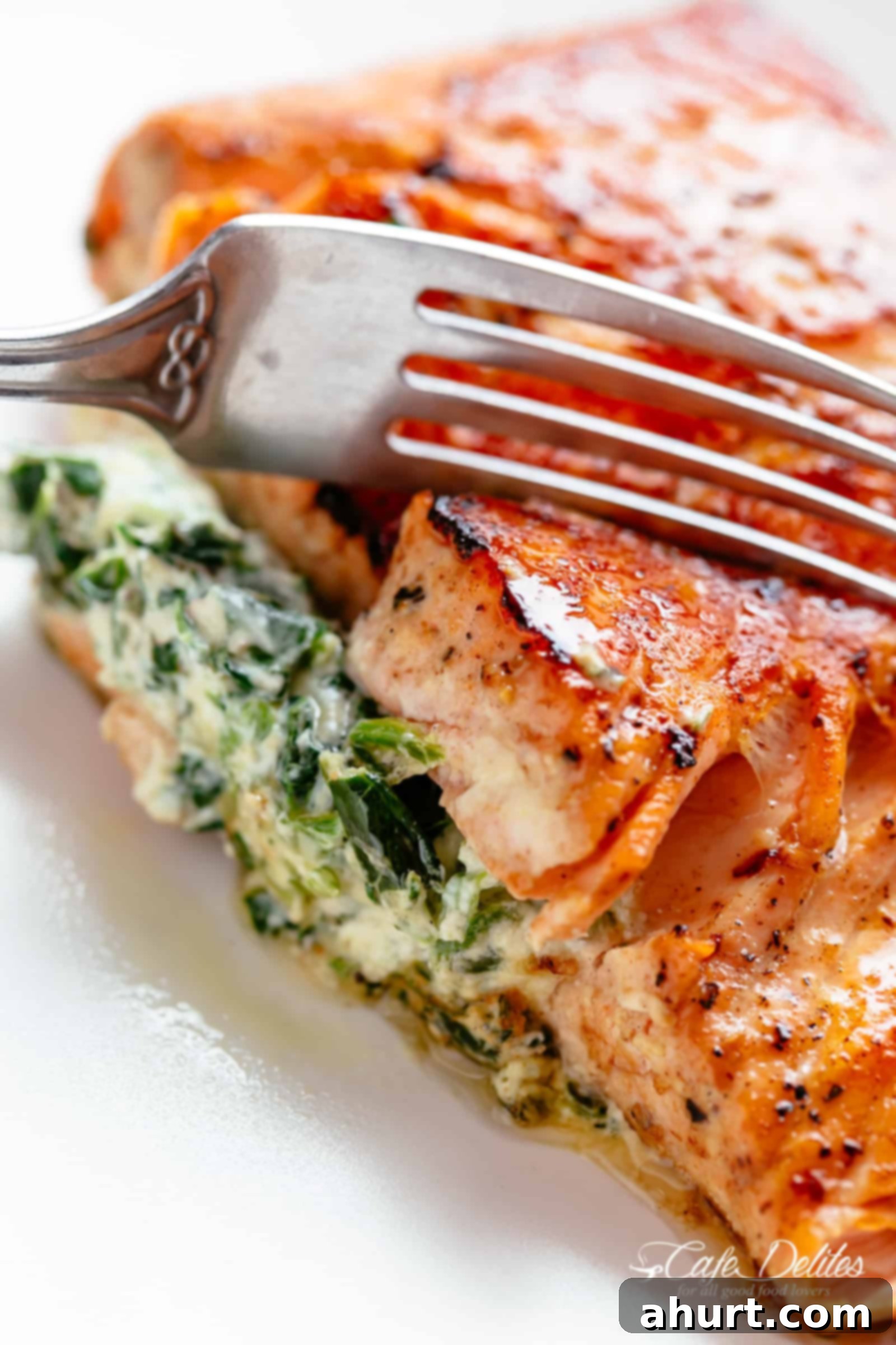 Decadent Garlic Butter Salmon Stuffed with Creamy Spinach 2 A fork slices through the side of a stuffed salmon fillet on a white plate. Filled with cream cheese, spinach, parmesan cheese and garlic.