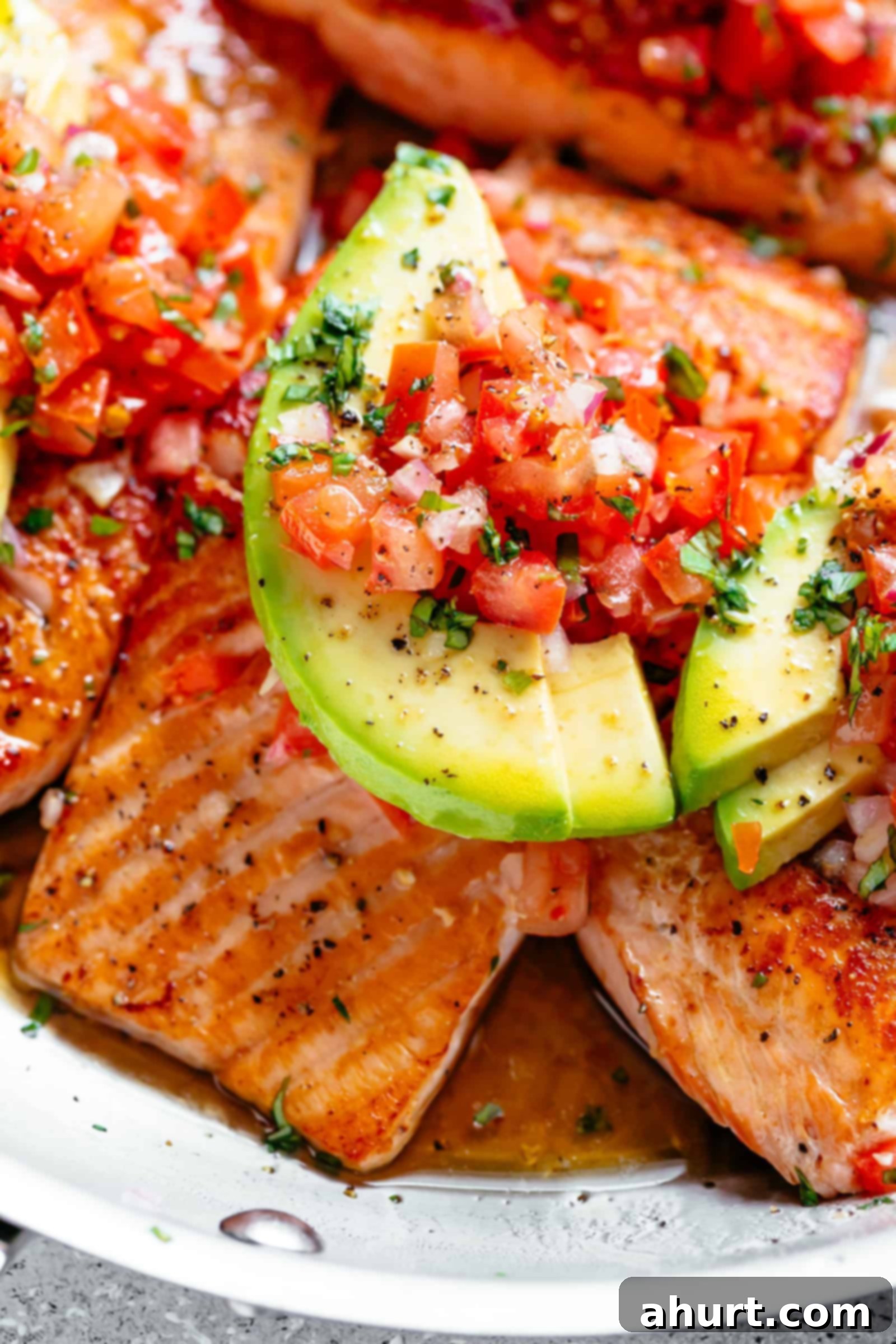 Close-up of three pieces of pan-seared salmon glistening in a buttery garlic lemon sauce, topped with fresh tomato salsa and avocado slices.