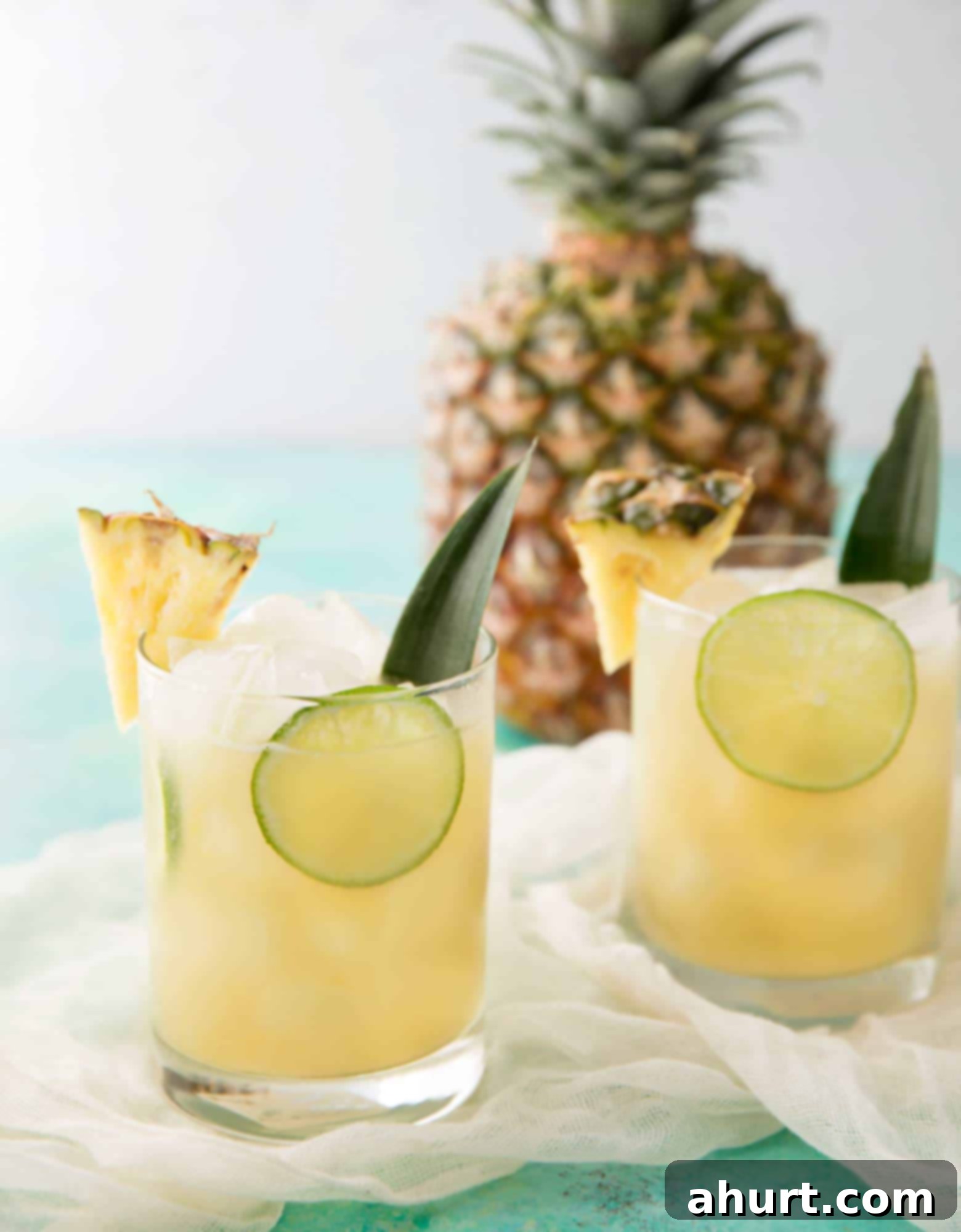 One sip of this tropical Pineapple Rum Punch and you'll be beachside in your mind! | cafedelites.com Two glasses filled with vibrant Pineapple Rum Punch, garnished with fresh pineapple and lime slices, set against a blurred tropical background.