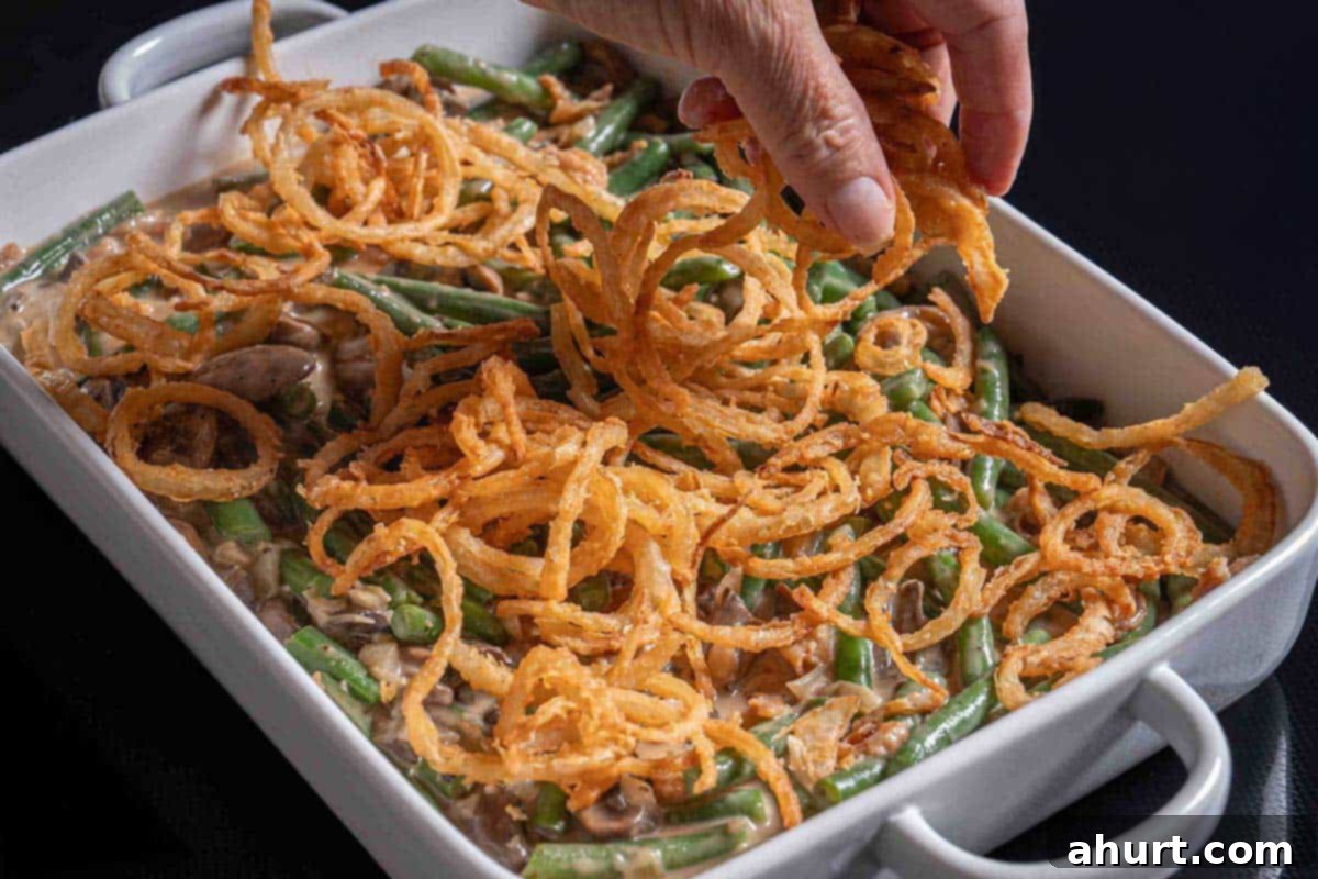 Velvety Green Bean Casserole with a Golden Crunch 10 green bean casserole in a baking dish being topper with fried onion rings