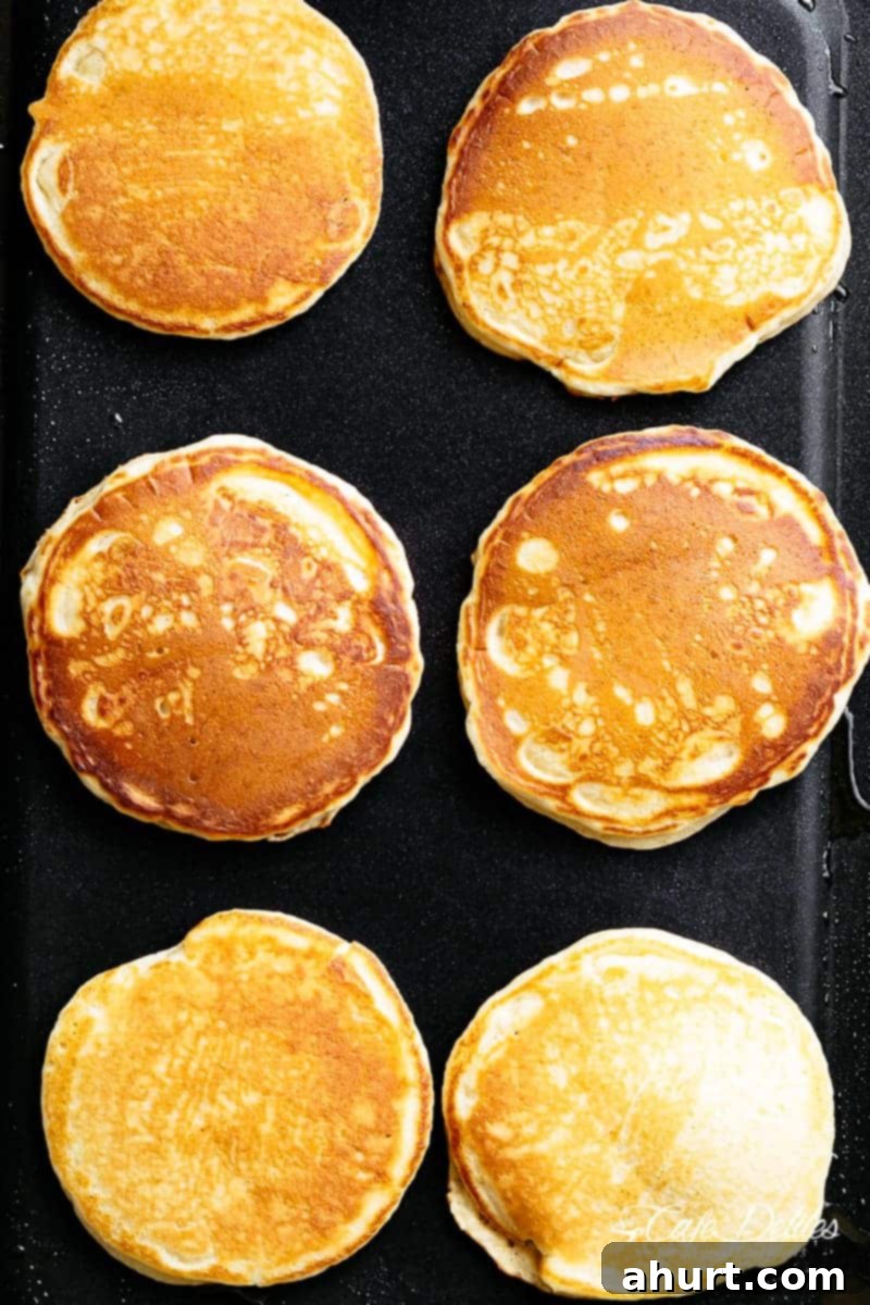 Two fluffy buttermilk pancakes cooking on a hot black non-stick griddle, showing bubbles forming on the surface before flipping.