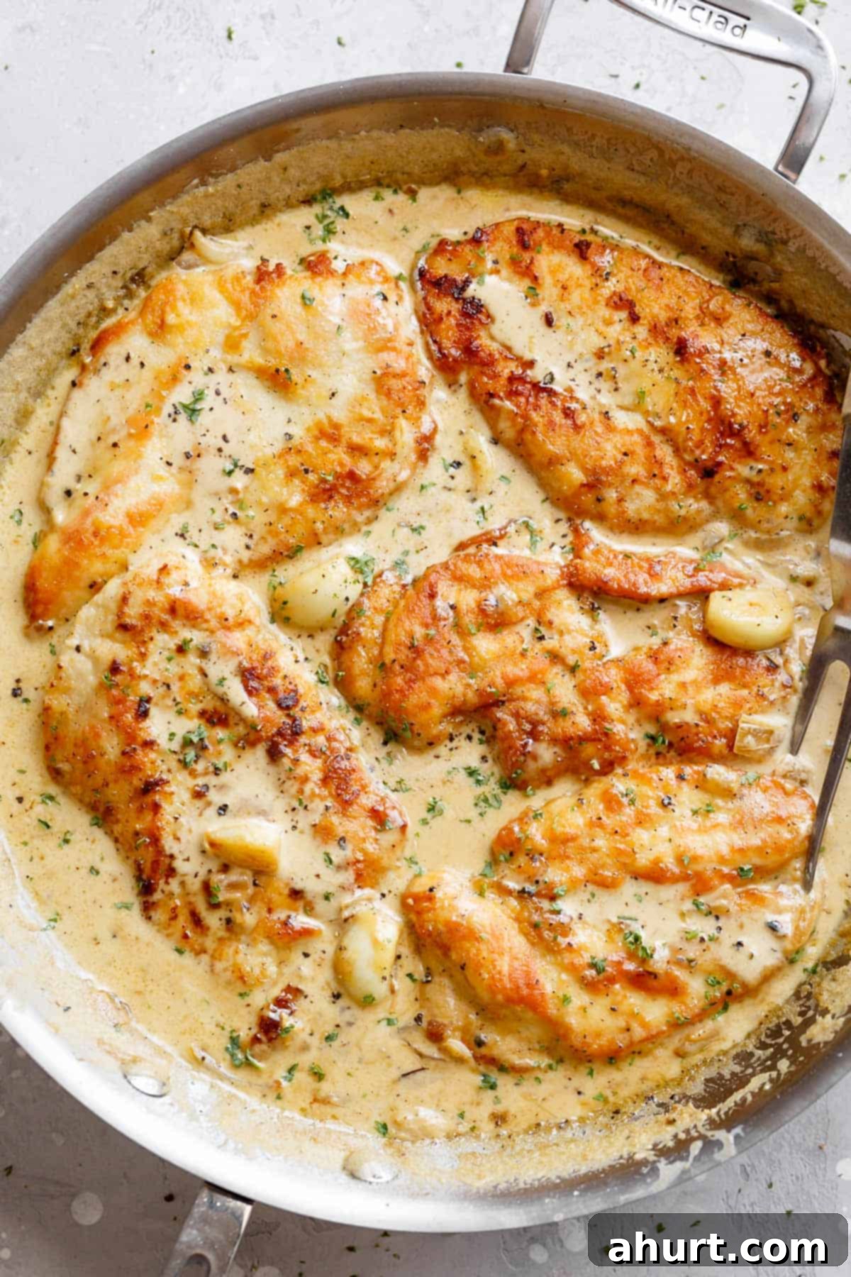 Irresistible Creamy Garlic Chicken Breasts 2 A silver frying pan filled with five beautifully cooked chicken breasts swimming in a luscious garlic cream sauce, garnished with whole roasted garlic cloves and a silver serving fork.