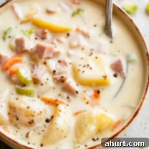 Rich Ham and Potato Chowder 9 creamy potato soup with a silver spoon resting in the top right corner. The bowl is on a marble bench top, a grey kitchen towel to the left of the bowl, and green celery bits scattered near the top of the frame.The soup has diced ham, potatoes, carrots, and is topped with pepper.
