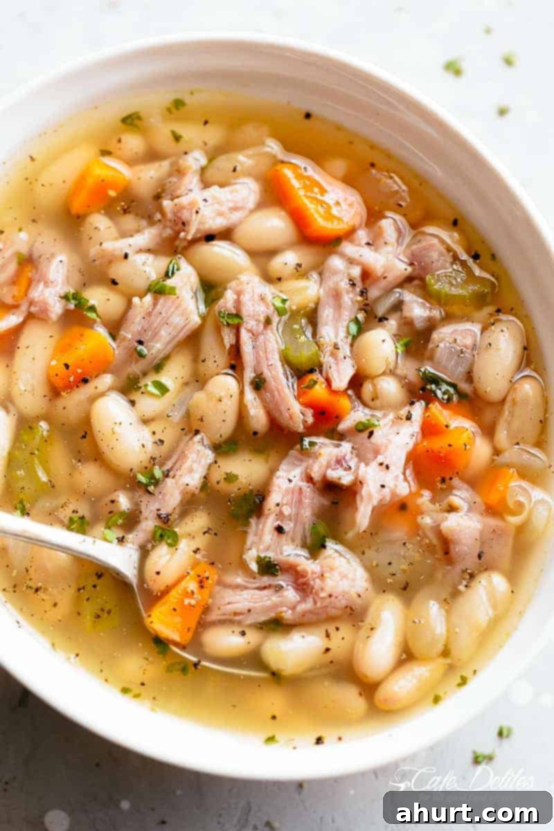 Smoky Ham and White Bean Chowder 2 Close-up image of a hearty ham and bean soup in a white bowl, with a spoonful being lifted, showcasing the tender beans and chunks of ham.
