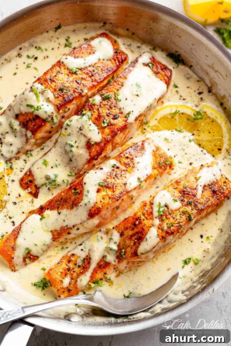 Decadent Garlic Butter Salmon 2 Four salmon fillets in a creamy garlic butter sauce in a large pan with a serving spoon resting inside the pan. Fresh green herbs are sprinkled over top of the fish and two lemon wedges sit beneath two of the fillets, looks yummy !