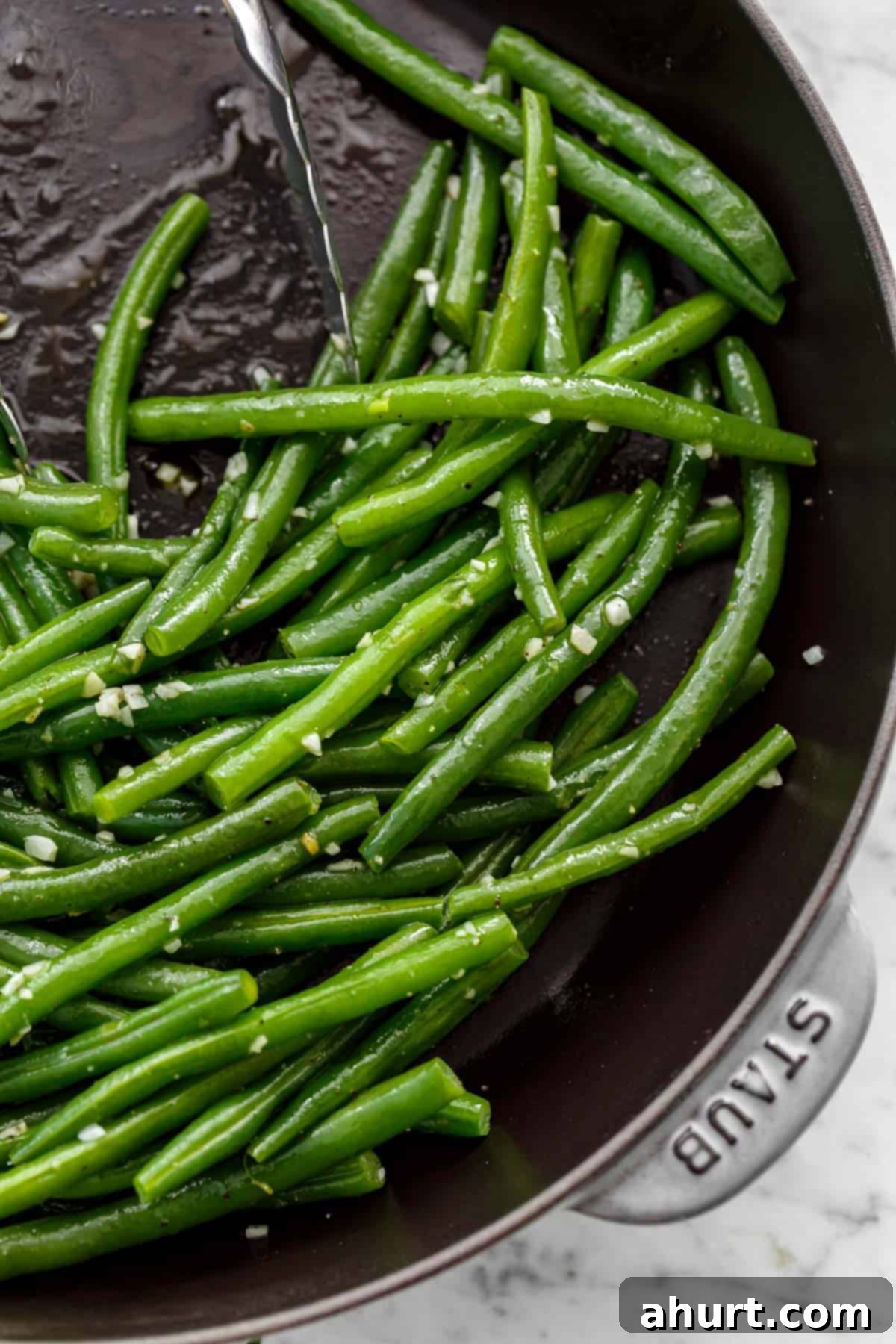 Close up image of Sautéed green beans with a sprinkle of course salt in a pan being pushed aside by a pair of tongs.