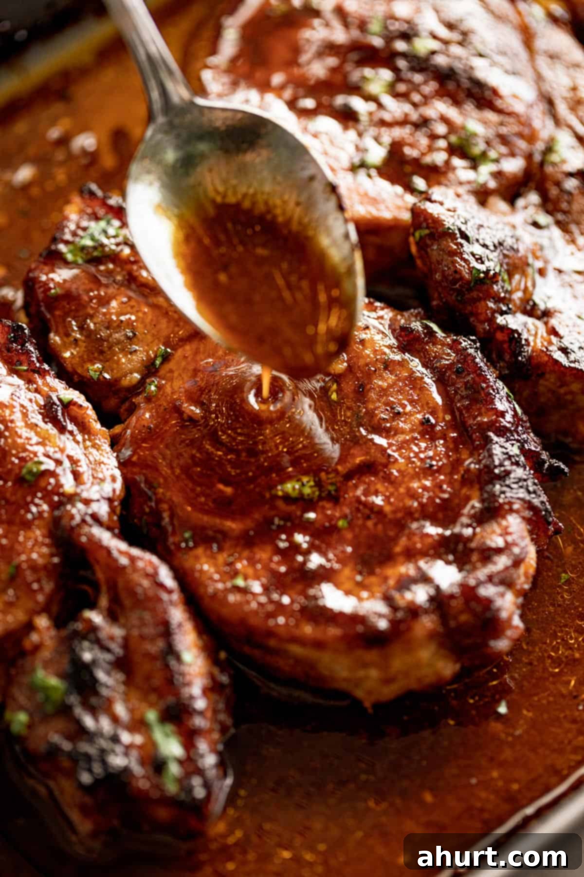 Tender Baked BBQ Pork Chops 3 Close-up image of a silver spoon pouring thin brown sauce over BBQ baked pork chops, highlighting the glistening finish.