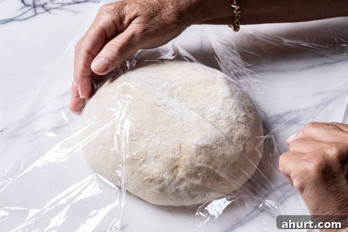 Effortless Artisan Crust: No-Knead Perfection 13 An image of hands covering the dough with parchment paper and then wrapping it in cling film.