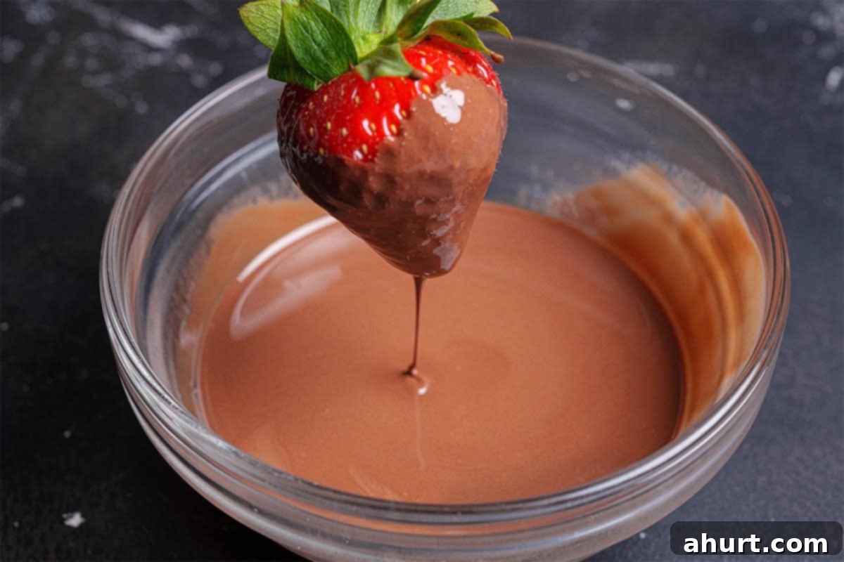 Tipsy Chocolate Strawberries 8 A single, perfectly dry strawberry being carefully dipped into a bowl of warm, melted chocolate using a fork, ensuring full coverage.