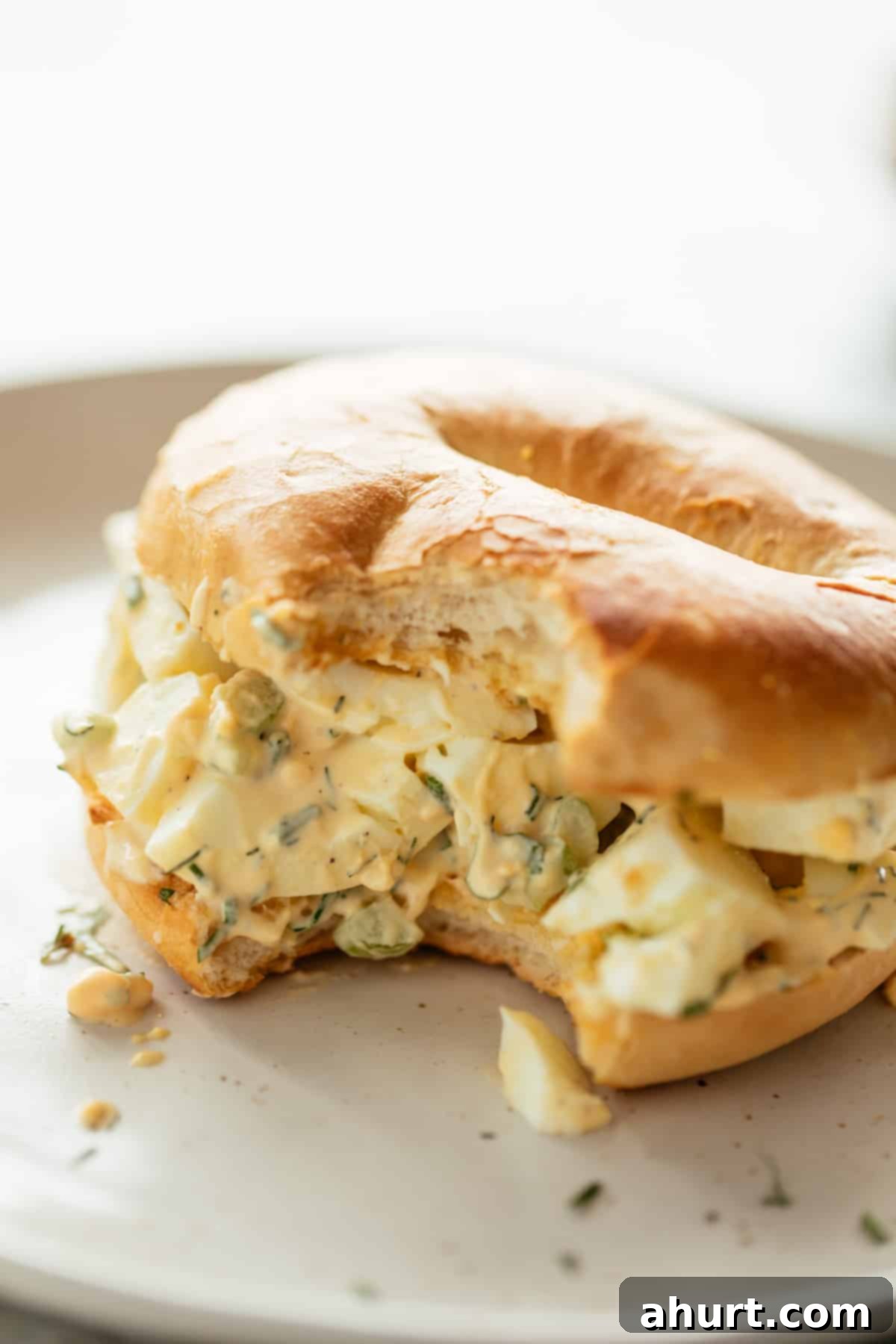 The Ultimate Egg Salad 10 Perfect Egg Salad sandwiched in a bagel on a white plate sprinkled with fresh dill, chives and parsley.