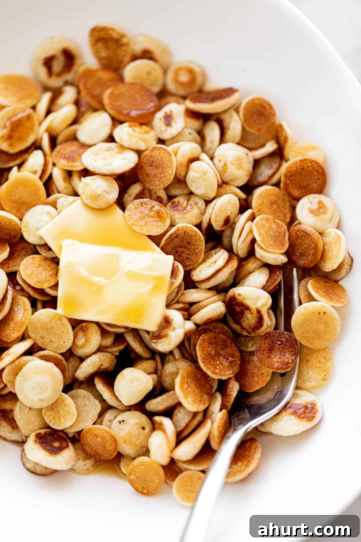 Mini Pancakes Cereal served in a white bowl with a silver fork. Topped with a piece of butter and drizzled with maple syrup.