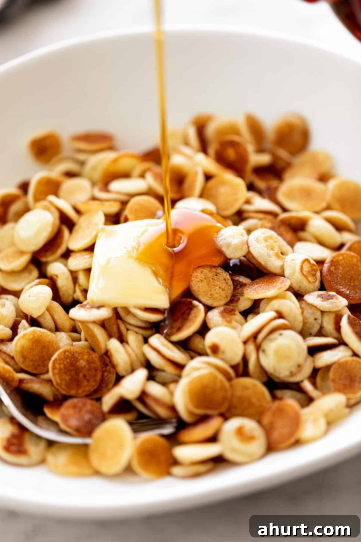 Maple syrup drizzled over Mini Pancakes Cereal served with a piece of butter in a white bowl with a silver fork.