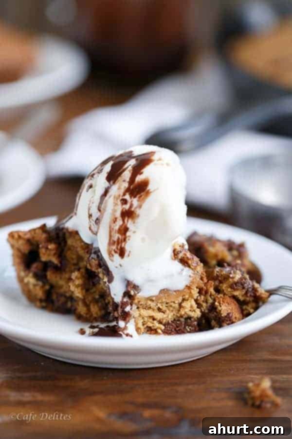 A close-up of a warm slice of Peanut Butter Chocolate Chip Deep Dish Skillet Cookie, topped with a scoop of melting vanilla ice cream, highlighting its soft interior.