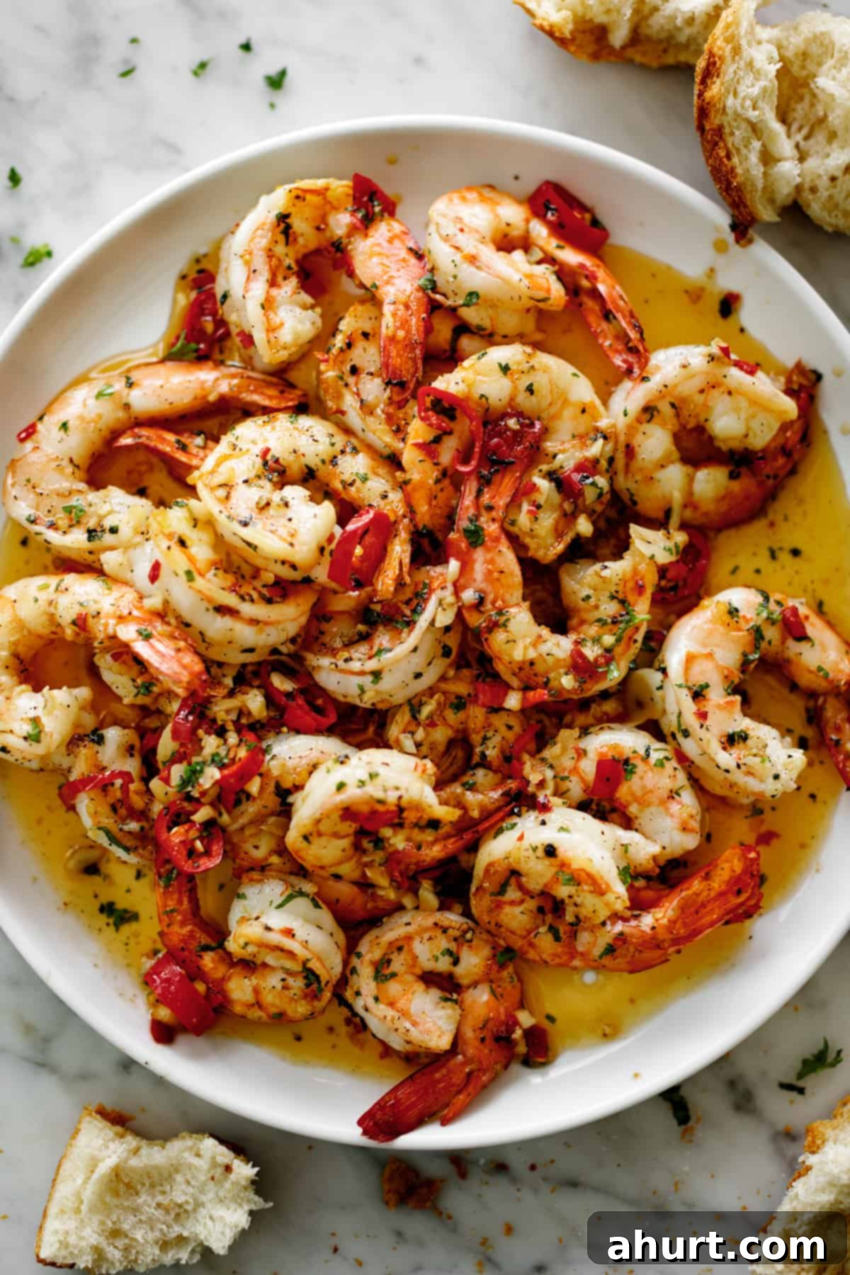 Perfect Garlic Butter Shrimp 2 A pile of succulent chili garlic butter shrimp on a white plate, coated in a vibrant, rich sauce with visible chili flakes and fresh parsley. Served with broken pieces of crusty bread, perfect for soaking up the delicious butter sauce.